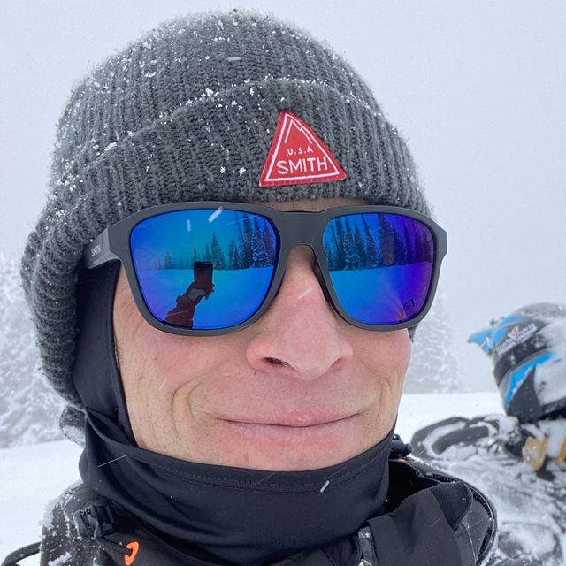 Guide Matt Hightower in a gray beanie and blue mirrored sunglasses, smiling in falling snow beside his snowmobile