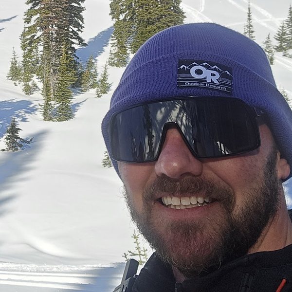 Guide James Clouse in a purple beanie and sunglasses in front of a snowy, tree-covered slope