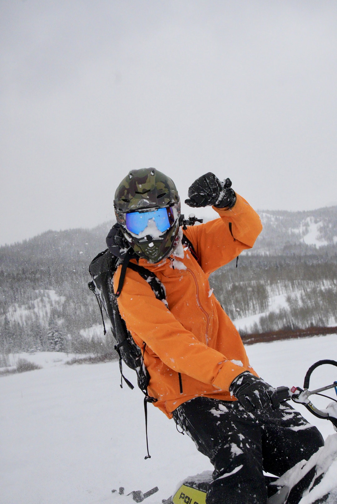 Guide John Spriggs in an orange jacket and camo helmet on a snowmobile in fresh powder
