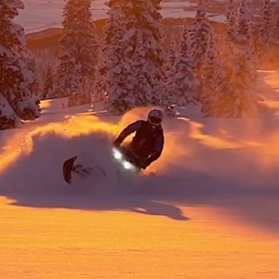 Guide Merlin Smith carving a snowmobile through deep powder at sunset