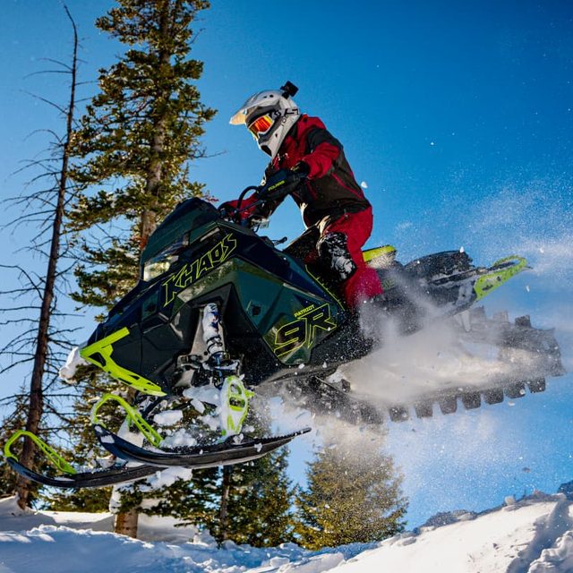 Guide Brandon Siok airborne on a green snowmobile in red gear against a blue sky